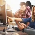 Closeup of group of application developers discussing about certain code for an application. There are two men and a woman and she's pointing at computer screen. Blurry people in background, also released. Orange light flaring from the background.